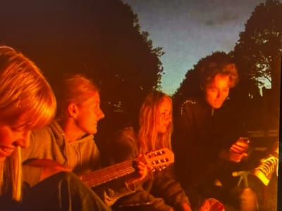 Four people are gathered around a fire at dusk in a wooded area. The subjects are illuminated by the firelight, casting a warm orange glow on their faces. One person on the left has blonde hair and is partially visible. Next to them, another person with blonde hair is playing a guitar. The guitar is light brown and has six strings. A third person with blonde hair is visible in the center, with their head turned to the side. The fourth person on the right, with curly dark hair, is looking down at their phone. They are wearing a dark top and striped shoes. The sky in the background is a dusky blue, with silhouetted trees against it. The scene suggests a casual gathering, possibly a camping trip or an outdoor hangout. The time of day is clearly evening, and the weather appears calm. No visible text is present in the image.