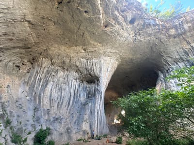 The image shows the Prohodna cave, also known as the "Eyes of God," located near the village of Karlukovo, Bulgaria. The cave is a large natural rock formation with two large openings that resemble eyes. In the foreground, there are several people at the base of the cave. One person is standing and looking up, possibly at a rock climber who is scaling the left side of the cave wall. Another person is lying down near climbing gear, which includes ropes and bags. The cave walls are made of gray rock with visible striations and vegetation growing on the lower sections. The opening of the cave leads into darkness, with some light filtering through from an unseen source further within. Green trees and foliage are visible on the right side of the image, framing the entrance to the cave and the sky above.