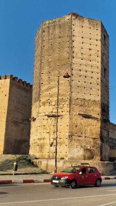 The image captures an outdoor urban scene in Mechouar Fes Jdid, Morocco, featuring historical fortifications and a contemporary vehicle. A tall, octagonal stone tower, constructed from light brown, weathered masonry, dominates the central-left. Its surface is characterized by numerous small, dark, recessed holes arranged in vertical and horizontal rows. A bird's nest is visible on the uppermost left corner of the tower. To the left and behind this prominent structure, additional sections of fortress walls, matching the tower's material and featuring crenellated tops, extend.
In the foreground, a paved road with a white solid line and a red-and-white painted curb runs horizontally. A red four-door sedan, identifiable as a taxi by a yellow rooftop sign, is in motion, moving from right to left on the road. The vehicle has a black front bumper. A dark, tall metal streetlight pole, topped with an ornate, dark brown luminaire, stands between the road and the tower. To the left of the road, a raised area with sparse green vegetation is visible, featuring a black and yellow cylindrical waste bin. The sky above is clear and bright blue. Long shadows are cast towards the left, indicating natural illumination from the right. Several small, dark avian figures are discernable against the sky and the fortress walls.