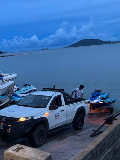 A man in a white t-shirt is leaning into the bed of a white pickup truck, which is parked on what appears to be a ramp or dock. The truck is loaded with what look like jet skis or personal watercraft. The scene is set at dusk, with a twilight sky and the ocean in the background. The water is calm and a dark, deep blue, reflecting the sky. A distant landmass, possibly an island or mountain range, is visible on the horizon. The overall mood is peaceful and serene, with a hint of activity as the man attends to the cargo. Visible text on the truck includes a "LIQUI MOLY" sticker.