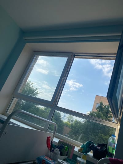 The image shows a view from a window, likely in a room with light blue walls. The window itself is large and appears to be a casement window, with the top section open, revealing a bright blue sky with scattered white clouds. Outside, green trees and a brick building are visible.
On the windowsill inside the room, several objects are present: a white plastic container holding a red toothbrush and other items, a packet of baby wipes, a bottle of green liquid, a bag of chips, and a plastic cup. A metal rail, possibly part of a bed or medical equipment, is in the foreground.
The scene is a close-up shot, primarily focusing on the window and the items on the sill. The lighting suggests it is daytime, with natural light streaming in. The location is identified as Chelyuskinsky, Russia.