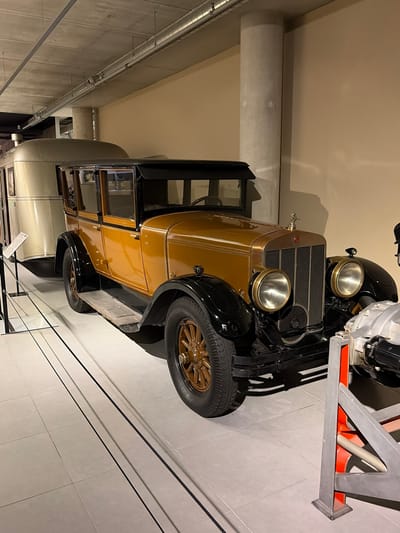 The image captures a vintage automobile displayed in an indoor exhibition space, likely a museum. The car, the primary subject, is a classic model from an earlier era, distinguished by its two-tone color scheme: a rich golden-bronze for the main body panels and contrasting black for the fenders, running boards, and roof. It features large, prominent circular headlights with metallic bezels, a vertical radiator grille, and multi-spoke wheels in a matching golden-bronze hue. A spare tire is mounted on the side of the car, and a small, intricate ornament (possibly a mascot) adorns the radiator cap.
The car is stationary, presented on a light-colored tiled floor. The background consists of plain, light-colored walls (beige or cream) and exposed ventilation ducts running along the ceiling or upper wall. To the right of the car, a metallic structure with silver and orange elements is visible, possibly a part of another exhibit or a display support. To the left, further into the background, part of another light-colored vehicle is discernible behind a glass barrier, suggesting multiple exhibits in the same space.
The scene is well-lit by artificial indoor lighting. No people are visible, emphasizing the static display of the exhibit. A small white sign or label can be seen near the glass barrier in the bottom left, but any text on it is illegible. The overall setting is clean and modern, providing a neutral backdrop that highlights the vintage vehicle.