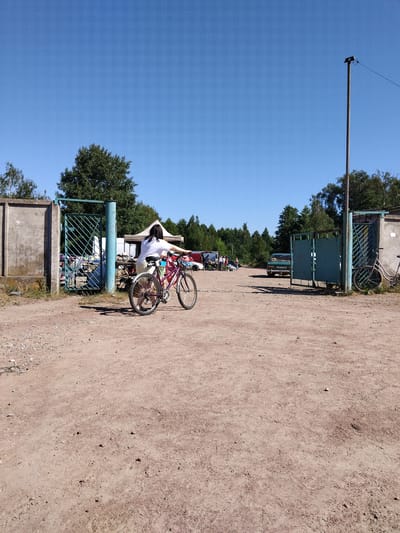 The image depicts a scene at an outdoor market or event with a young woman on a pink bicycle in the foreground. She is wearing a white top and light-colored shorts, with her back to the camera, facing towards an open gate. The bicycle is positioned on a dirt path. Behind her, through the green metal gate, appears to be a bustling market with tents, tables, and various goods displayed. Several vehicles, including a green car with a license plate that reads "M 18 49 BH", are visible in the background, along with other people browsing the stalls. The environment is outdoors, with a clear blue sky and trees lining the perimeter of the market area. A tall pole with some electrical equipment is situated to the right of the frame. The overall setting suggests a sunny day at a local marketplace in Irshansk, Ukraine.