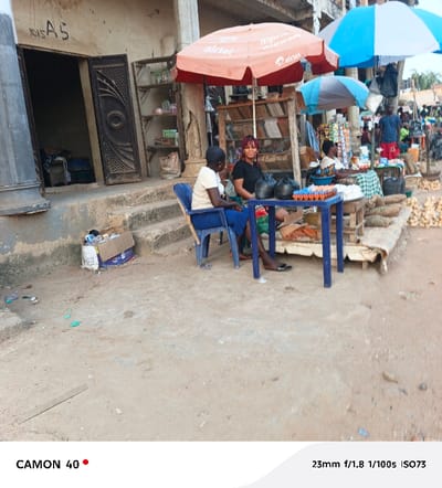 A street scene shows two women seated at a blue table under an orange umbrella labeled "airtel." The table displays eggs, with produce laid out behind it. Behind them, more vendors and pedestrians are visible. To the left, a partially open, ornate door leads into a building, next to a set of steps where a cardboard box sits. The ground is dusty and uneven.