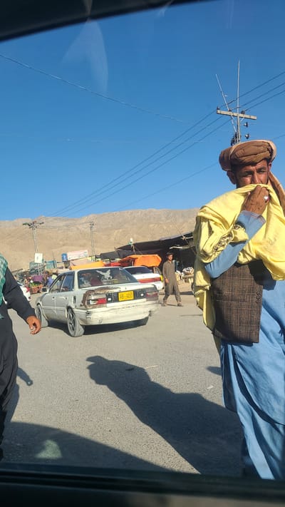 The image depicts a street scene, presumably taken from inside a vehicle, in what is likely Kuchlak, Pakistan, based on the license plate format and surrounding environment. An elderly man dressed in traditional clothing, including a turban and a yellow and blue robe with a brown waistcoat, stands in the foreground. He appears to be speaking to someone, perhaps the photographer.

The background shows a street with parked cars, notably a white vehicle with the license plate "AB-4233". A small market or roadside shops are visible along the street, and a man is walking along the roadside. Utility poles and wires are visible against a clear blue sky. The terrain in the distance is mountainous and arid.

The lower portion of the image contains a shadowed arm and part of a person's clothing, likely the photographer. The overall setting suggests a small town or semi-urban environment within a mountainous region.