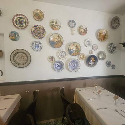 The image depicts an interior scene of a restaurant, likely in Lloret de Mar, Spain, as suggested by the decorative plates adorning the walls. The walls are painted a light, textured color and are covered with a collection of ceramic plates, many featuring colorful, intricate designs of landscapes, people, and geometric patterns. Some plates show scenes of rural life, villages, and nautical themes, characteristic of Mediterranean artistry.
Below the plates, a dark brown wainscoting runs along the wall. In the foreground, two tables are set for a meal. Each table is covered with a white tablecloth and is laid with cutlery (forks and spoons) and glasses with folded napkins. The presence of dining tables and place settings indicates that the restaurant is either open for business or prepared to welcome customers.
The lighting suggests it could be daytime, possibly late afternoon or early evening, given the indoor setting. The atmosphere appears to be calm and relaxed, with the focus on the decorative elements of the restaurant's interior. No people are visible in the image, and there is no discernible text. The overall impression is that of a charming and traditional dining establishment.