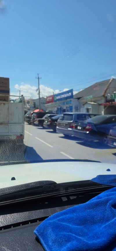 The image captures a street scene in Karakol City, Kyrgyzstan, on a bright, sunny day. The perspective is from inside a vehicle, looking out through the windshield. Parked cars line the right side of the road, which is paved and marked with white lines. The foreground shows the interior of the vehicle, including the dashboard and a blue microfiber cloth.
In the middle ground, various vehicles are visible. On the left, a white truck, possibly a cargo truck, is partially visible. To its right, several cars are parked, including dark sedans and SUVs. The buildings lining the street appear to be commercial establishments, with signs visible. One prominent blue sign with white text that appears to say "SAFE DEPOSIT" or similar can be seen on a building. Another red sign is also present. Power lines and poles are visible against the clear blue sky, which has some scattered white clouds. The overall impression is that of a typical urban street, possibly in a commercial district, with moderate traffic and parking.