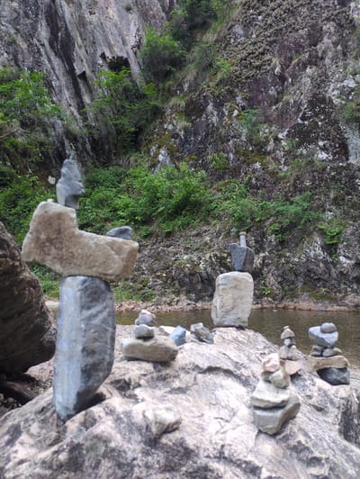 This image depicts a natural outdoor scene in Godang-ri, South Korea, characterized by a steep, vegetated rock face and a body of water.

In the immediate foreground, a large, irregularly shaped rock surface occupies the lower half of the frame. This rock is predominantly light gray with darker striations and patches. Positioned on this surface are multiple cairns, or stacks of balanced stones, varying in size and composition. The most prominent cairn is located on the left, constructed from a dark gray, elongated base stone, supporting a lighter, broad, horizontal stone, which in turn supports several smaller, dark gray stones at its apex. To the right of this, several smaller cairns are visible, typically comprising three to five irregularly shaped stones stacked vertically. One notable cairn in the mid-right consists of a light-colored base stone topped by a dark, somewhat bottle-shaped stone, further topped by a smaller, rounded stone.

In the midground, a narrow, calm body of water, likely a river or stream, extends horizontally across the frame. The water exhibits a murky, brownish-green hue, with reflections visible on its surface. The banks appear to be composed of small stones and earth.

The background is dominated by a very steep, massive rock cliff face. This cliff displays varied textures, with prominent vertical and diagonal fissures and crevices. The rock itself is a mix of dark gray, brown, and lighter weathered patches. Abundant vegetation covers large portions of the cliff; this includes dense clusters of bright green bushes and smaller trees primarily at the base and in the lower crevices. Higher up, patches of light green moss, lichen, and scattered tufts of dry, straw-colored grass cling to the rock surface. The overall lighting suggests daylight, with no harsh shadows, indicating potentially overcast conditions. No people or animals are visible in the scene.
