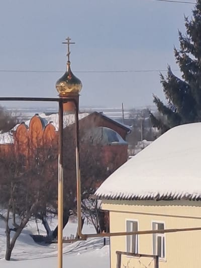 The image displays a winter scene in Bolshaya Elhovka, Russia, under a clear, bright blue sky. Centrally, a golden onion dome, topped with a distinct Orthodox cross, is visible, supported by a vertical metal framework. To the left and slightly behind this structure, a large, reddish-brown building with multiple snow-covered, rounded roof segments is present. The foreground and midground are covered in a uniform layer of snow, with several bare deciduous trees scattered throughout the snowy terrain. On the right side of the frame, a portion of a light yellow building with two visible windows is depicted; its steeply pitched roof is heavily laden with snow, forming prominent icicle-like overhangs along the eaves. An evergreen tree is partially visible on the far right. Overhead power lines horizontally traverse the upper section of the image. The distant background consists of a flat, snow-covered expanse. No individuals or active interactions are observed.