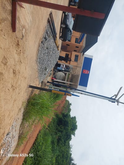 The image depicts a construction site and a fuel station in Ogbunike, Nigeria. In the foreground, a pile of gravel is visible next to a sandy area. Several large pipes, likely part of the construction, are laid out on the ground. Further back, a fuel station with a distinctive blue and white structure stands, featuring the logo of "OANDO". A black car is parked near the station, and a few individuals are present, some appearing to be working. The background shows buildings and lush greenery, indicative of the tropical environment. The sky is overcast. The image also displays a watermark in the lower-left corner: "Galaxy A52s 5G".