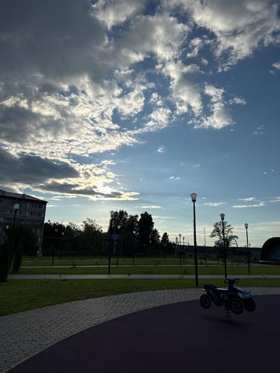 The image depicts a playground area in an urban setting, possibly a park or residential complex, with a clear sky and scattered clouds. In the foreground, a purple rubberized surface is visible, transitioning to a paved walkway made of grey interlocking bricks. A dark blue spring-mounted toy vehicle, resembling an ATV, is positioned on the purple surface.

The background features a manicured lawn with several streetlights. Beyond the lawn, a line of trees forms a dense green barrier. To the left, a multi-story apartment building with a gray facade is visible. The overall lighting suggests late afternoon or early evening, with sunlight breaking through the clouds. The scene appears calm and devoid of human activity. The location is identified as Mozhga, Russia.