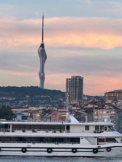 The image shows a view of the Çamlıca Tower and a modern multi-story building in Kadıköy, Turkey, set against a sky with wispy pink and orange clouds. In the foreground, a white, multi-deck ferry or cruise ship is moving across the water, with several people visible on its decks. The ship has a red waterline and black tires mounted along its sides. Behind the ship, a dense urban landscape of residential buildings with red-tiled roofs and varying architectural styles fills the midground. The Çamlıca Tower, a prominent landmark, is visible in the distance, towering over a tree-covered hill. The overall scene depicts a coastal urban environment during either sunrise or sunset.