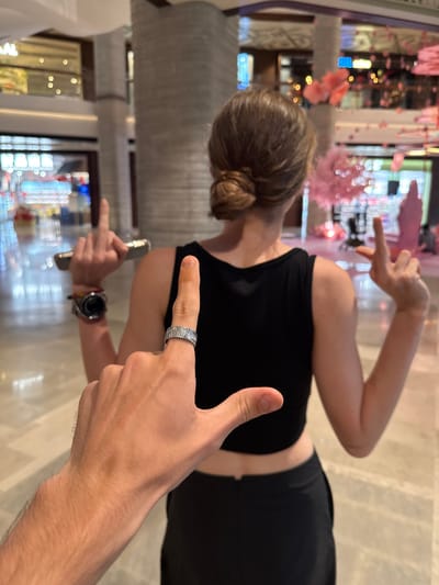 The image, originally rotated 90 degrees counter-clockwise, depicts an indoor scene likely within a commercial complex or shopping mall in Putuo District, China.

In the immediate foreground, the right arm and hand of one person are prominently featured. Their index finger points directly towards another individual, and a distinctive silver-toned ring with a textured, wavy pattern adorns this finger. The foreground person's lower chest and midriff, clad in a black top, are also partially visible.

In the mid-ground, a second individual is seen from behind and slightly to the side, with their head turned away from the camera. This person has light brown hair styled in a neat bun and is wearing a black sleeveless or crop top and dark bottoms. They are actively making a dynamic hand gesture with both hands, fingers splayed outwards. A watch is visible on their left wrist, and they hold a silver-colored smartphone or similar device in their left hand, also pointing with their index finger.

The background is intentionally blurred, creating a bokeh effect that highlights the subjects. Despite the blur, elements consistent with a modern, well-lit interior are visible. Pink, floral-like decorations are suspended or arranged throughout the space, possibly as part of a seasonal display or an artistic installation. Glimpses of illuminated storefronts or displays with various lights can be seen in the soft-focus distance, suggesting a bustling commercial environment. The floor is a light-colored, reflective surface, possibly tiled or polished concrete.

The activity appears to be a casual or playful interaction, possibly a posed photo op, characterized by the hand gestures and pointing. The lighting suggests a bright indoor setting, whether daytime or a well-illuminated evening. There is no discernible text visible in the image.