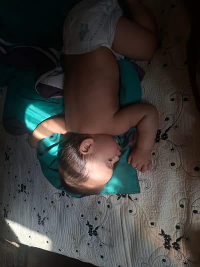 A baby is sleeping on a bed, angled on its left side, with its head resting on a teal pillow and its body covered partially by a teal fabric. The baby is wearing a diaper. Sunlight streams across the baby's arm and chest, creating a pattern of light and shadow. The bed appears to have a white quilted blanket with a floral pattern in black. The image is oriented with the baby's head to the left and feet to the right, rotated 90 degrees counter-clockwise. The scene is indoors, in a residential setting within the city of Tomilino, Russia.