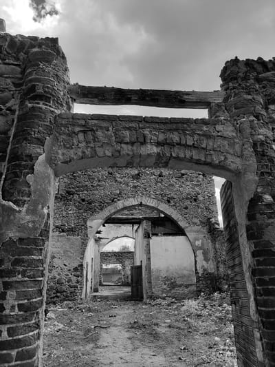 The image shows a partially ruined brick building in black and white. The building's walls are crumbling, revealing the brickwork and stone construction. There are arched openings and doorways leading into the interior of the structure. The sky is visible through openings in the building. The location is Жалудоцкі сельскі Савет, Belarus. The scene is static, depicting the state of disrepair and abandonment of the building.