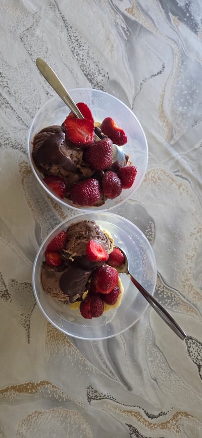 Two bowls of chocolate and vanilla ice cream topped with strawberries sit on a marble-patterned surface. Each bowl is made of clear plastic and contains scoops of ice cream, some of which are melting. Whole and sliced strawberries are scattered on top of the ice cream. A spoon is visible in each bowl, one scooping a strawberry and the other resting in the ice cream. The lighting suggests it is daytime, and the scene is likely indoors, given the controlled lighting and setting. There is no visible text in the image.