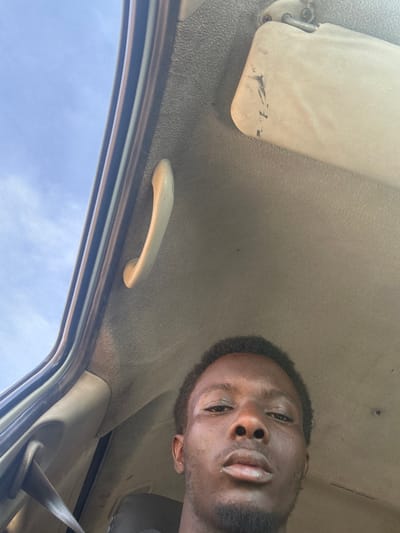 The image depicts a low-angle perspective of a young adult male positioned inside a vehicle, presented as being located in Aboro, Nigeria. The male subject, appearing to be of African descent, occupies the lower half of the frame, looking directly at the camera with a neutral expression. He has short, dark, curly hair and a short, neatly trimmed beard.
Above the subject, the vehicle's interior ceiling is visible, upholstered in a light gray, textured material that shows some discoloration and minor wear. A beige-colored grab handle is affixed to the ceiling panel, positioned to the upper left of the subject. Further to the right in the upper frame, a beige sun visor is present, displaying dark smudges and signs of wear along its lower edge.
On the far left, a portion of the vehicle's dark-colored window frame or door pillar is visible, outlining an opening through which a clear, light blue sky is discernible, suggesting an outdoor setting or an open window. A dark-colored seatbelt strap is also partially visible in the lower left corner, near the subject's left shoulder. The overall scene suggests a selfie taken from the driver or passenger seat of an older model vehicle.