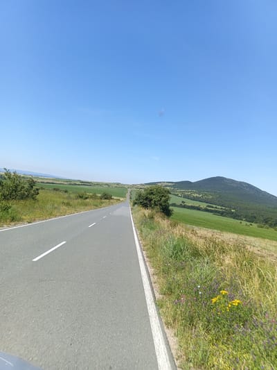 The image shows a road with a white dividing line, stretching into the distance and curving to the right. The road is bordered by a grassy shoulder with wildflowers. To the right of the road, there is a lush green field with a conical hill covered in trees in the background. To the left of the road, there are more green fields and scattered trees. The sky is clear and blue. The scene appears to be a rural landscape in the vicinity of Krumovo, Bulgaria, likely during daytime. There are no people or animals visible in the frame.
