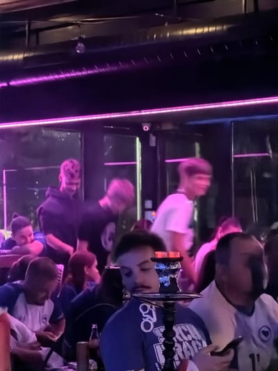 A group of young adults are gathered in a dimly lit indoor venue, possibly a hookah lounge or club, in Travnik, Bosnia and Herzegovina. The atmosphere is characterized by vibrant pink neon lighting that illuminates the space. In the foreground, a hookah pipe is prominently displayed, with glowing charcoal on top. Several individuals are visible, some engaged in conversation and others looking at their phones. The people appear to be in their late teens or early twenties, dressed casually. The background reveals more of the venue's interior, including tables and other patrons, all bathed in the purple glow of the lights.