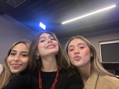 Three young women pose closely together, their faces tilted towards the camera in an overhead shot. One woman wears a tan jacket, another a black top with a red necklace, and the third has a bare shoulder and a similar blonde hair color. A rectangular window is visible in the upper left, and a long, linear light fixture illuminates the scene below. The overall lighting is somewhat dim with direct lighting under the women's faces.