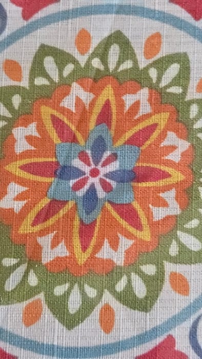 The image presents a detailed close-up of a patterned textile surface, originating from Lompoc, United States. The primary subject is a colorful, symmetrical floral or mandala-like motif centrally positioned. This motif is rendered with a prominent orange outer layer of stylized petals, outlined in yellow, surrounding an inner layer of red petals. The core of the design features blue, tear-shaped petals encircling a small, central white asterisk-like element with eight points, which itself is contained within a red circle. Surrounding the main orange and red floral structure are green, stylized leaf or petal shapes and small orange circular or oval elements, all set against a textured, light off-white woven fabric background. The visible coarse weave of the fabric reveals distinct horizontal and vertical threads throughout the entire frame. No individuals, explicit actions, or interactions are depicted. The immediate environment is solely the described fabric material.