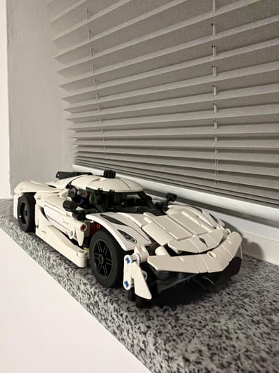 A white Lego Technic replica of a sports car is displayed on a speckled gray countertop. The car, facing right, is positioned in front of a set of closed, off-white horizontal blinds that cover a window. The background consists of a plain white wall to the left of the blinds. There are no people or animals present in the image. The scene appears to be indoors.
