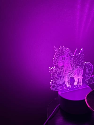 This image shows a close-up of a purple LED night light shaped like a unicorn, casting a vibrant purple glow onto a textured wall. The unicorn design is etched into clear acrylic and features intricate details, including stars, flowing mane, and a detailed depiction of the unicorn's head and upper body. The light source appears to be housed in a white cylindrical base, partially visible on the right side of the frame, with a dark shadow cast on the wall behind it. A thin white cord extends from the base, suggesting it is plugged into a power source. The overall scene is dark, with the light and its projection being the primary illumination. The purple hue dominates the image, creating a magical and whimsical atmosphere. The textured wall, lit by the light, shows subtle patterns and shadows, indicating it might be a room interior. No people are visible in the image. The location context provided is Peyia, Cyprus, but there are no specific visual cues in the image to confirm this. The image captures a decorative lighting element in use, likely in a bedroom or nursery.