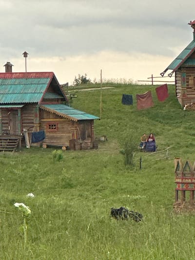 The image depicts a rural scene in Tokkari-Lend, Russia, featuring traditional wooden houses with turquoise roofs. The setting appears to be an outdoor historical reenactment or a village festival.

In the foreground, to the left, is a partially visible wooden structure with a turquoise roof and red trim. Laundry, including a red cloth, a brown cloth, and a dark blue cloth, is hung on a clothesline.

Further into the scene, a larger wooden house with intricate detailing and a red-framed window is visible. A dark blue tarp or blanket is draped over a wooden beam near the house.

In the middle ground, two people are sitting or lying on the grass. One appears to be wearing a blue garment, and the other, a lighter colored one. They are near a simple metal structure, possibly part of a game or activity.

To the right, a pile of dark objects, possibly part of a display or historical exhibit, is visible on the grass.

In the far distance, a brightly colored, patterned structure can be seen, suggesting a decorative element or another building.

The ground is covered in green grass, and the sky is overcast, suggesting a cloudy day. There is no visible text in the image. The overall impression is one of a rustic, historical, or cultural gathering in a rural Russian setting.