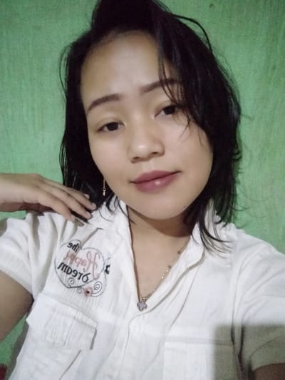 A young woman poses in a selfie against a green wall. She has dark hair and brown eyes, with a neutral expression. She wears a white, button-down shirt with an embroidered design on the upper left side. A delicate necklace with a light purple pendant adorns her neck. Her makeup includes lipstick with a mauve tint. Her hand rests on her collarbone, adding a casual touch to the image.
