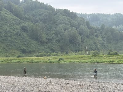The image depicts a wide river in Ust-Kabirza, Russia, with a steep, densely forested hillside rising on the far bank. The weather appears overcast, with a visible haze and small white specks, possibly indicating rain or pollen. In the foreground, a rocky shoreline borders the river. Two individuals are present in the water, each engaged in fishing. One person, on the left side of the frame, stands in waders and appears to be holding a fishing rod. The second individual, further to the right, also stands in the river, facing away from the camera, and is holding a fishing rod. A single utility pole with two crossbars is visible on the grassy bank behind the river, to the right of center. The overall scene suggests a tranquil, natural environment.