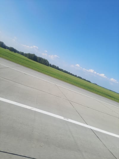 An outdoor, diagonal shot shows a paved surface with white markings, possibly a road or runway, that extends into the distance. To the right of the paved surface, a vibrant green field stretches to a line of trees in the background. Above the field and trees, a clear blue sky with a few wispy clouds dominates the upper portion of the image. The scene is likely located in Pokrovka, Russia, given the rural setting and vegetation.