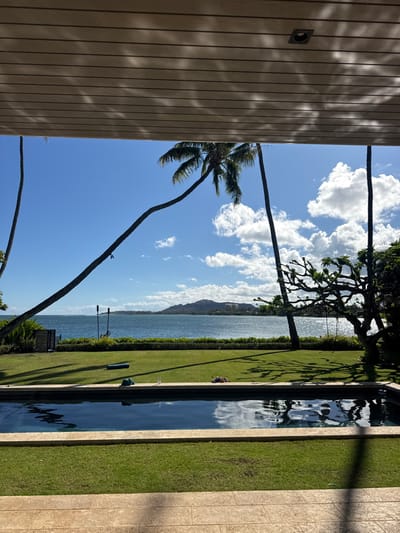The image displays a coastal residential property in East Honolulu, United States, viewed from an outdoor covered patio. The overhead structure is a light-colored, louvered ceiling with light patterns cast upon it. In the immediate foreground, a tiled patio floor is visible. An infinity-edge swimming pool with dark blue water occupies the central foreground, reflecting the sky and clouds.
Beyond the pool, a meticulously maintained green lawn extends to a low hedge bordering a body of water, identified as a bay or ocean. On the left side of the lawn, a dark rectangular mat and a blue object are present. Near the right edge of the pool, a clear glass or bottle stands upright. A pile of dark fabric or items is situated on the right side of the lawn.
Several trees are prominent: a tall, curved palm tree leans across the left side of the frame, a straighter palm tree stands further right, and a gnarled tree with dark, intricate branches is on the far right. Two dark, vertical poles, resembling tiki torches, are visible at the water's edge on the left.
The background features a large expanse of water, reflecting sunlight. Across the water, the distinct profile of Diamond Head (Lēʻahi) crater is clearly visible under a bright blue sky with scattered white cumulus clouds. No individuals are present in the scene. The environment indicates a sunny daytime.