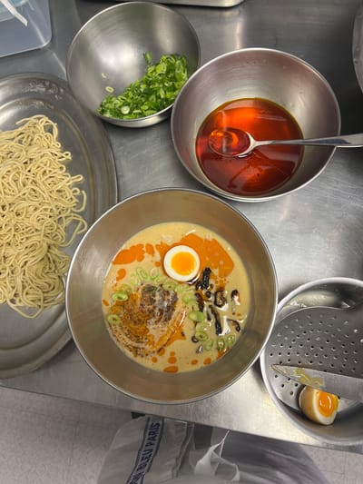 The image displays a professional kitchen or food preparation area, with the specific location in Pozuelo de Alarcón, Spain, not identifiable from the visual content. The scene focuses on the preparation and assembly of ramen.
In the center, a stainless steel bowl contains an assembled ramen dish. The dish features a creamy, light-colored broth with visible swirls of reddish-orange chili oil or flavored oil. Garnishes include sliced green onions, dark fungal elements resembling mushrooms, and a central piece of meat or paste topped with seasoning. A half-cut, soft-boiled egg with a firm white and a vibrant orange, runny yolk is positioned on the right side of the bowl.
To the upper left, a smaller stainless steel bowl holds a quantity of chopped green onions. Adjacent to this, on a flat, oval-shaped metal tray, a pile of uncooked or par-cooked ramen noodles is arranged. In the upper right quadrant, another stainless steel bowl contains a pool of reddish-orange oil, with a silver spoon resting submerged in it. In the bottom right, a perforated stainless steel strainer or bowl holds another half of a soft-boiled egg, which is being separated or cut by a silver knife or spatula.
The entire arrangement rests on a stainless steel countertop, exhibiting minor reflections and signs of a working surface. A white and blue striped cloth or towel, partially visible in the bottom center, bears text including "DU BLEU PARIS". A translucent plastic container is partially visible in the upper left background. No individuals are depicted, but their presence is implied by the organized food preparation.