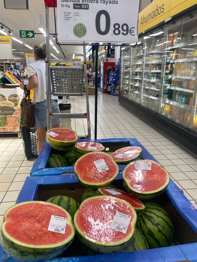 The image depicts the interior of a supermarket in Bel Air, Spain. In the foreground, several watermelons are displayed in a blue cardboard crate. Some are whole, while others are halved and wrapped in plastic. Price tags are visible on some of the watermelons. A price sign is also visible in the upper portion of the image, indicating "Sandia entera rayada" (striped whole watermelon) priced at 0.89€. In the background, a woman wearing a white t-shirt and denim shorts is seen from behind, pushing a shopping cart. Refrigerated display cases filled with various food items are visible along the right side of the image. The floor is tiled with white squares. An illuminated green emergency exit sign is visible on the left side of the background.