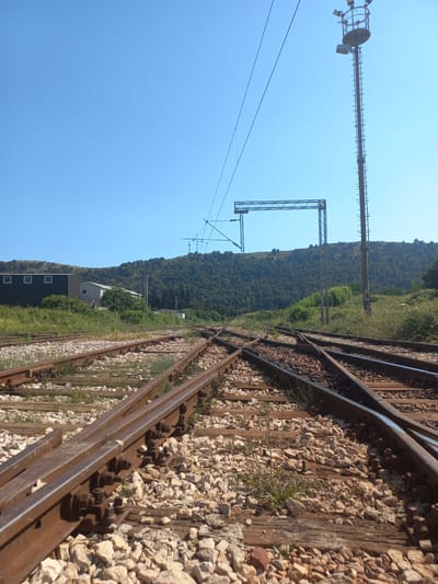 This media file captures a view of a railway track with a mountainous backdrop under a clear blue sky, suggesting a sunny day. The perspective is close to the ground, focusing on the railway infrastructure.

The primary subjects are the train tracks themselves, characterized by weathered metal rails and a ballast of light-colored stones. Wooden sleepers are visible beneath the rails, some showing signs of age. The tracks appear to be a junction, with multiple sets of rails diverging. To the left of the tracks, tall metal poles with overhead electrical wires, typical of electrified railways, are prominent. One such pole has a large circular apparatus and several lights attached, possibly a signal or a lighting fixture. Further in the distance, another metal structure resembling a gantry or support for overhead wires stands against the sky.

Beyond the railway, a steep, tree-covered hillside dominates the background. The foliage suggests a temperate or Mediterranean climate. Closer to the tracks, a low-rise building with a dark facade and several square windows is visible. Beside it, a lighter-colored, more modern structure with a sloped roof is also present. Green vegetation, including grass and bushes, grows alongside and between the tracks, indicating a natural, possibly rural or semi-rural, setting.

No people are visible in the frame, and no specific activity or event is clearly taking place. The overall impression is one of infrastructure within a natural landscape. The lighting suggests it is daytime, likely morning or afternoon, given the brightness and the clear sky. There is no visible text in the image. The location context provided is Polje, Montenegro, which aligns with the hilly, vegetated terrain and the presence of railway infrastructure.