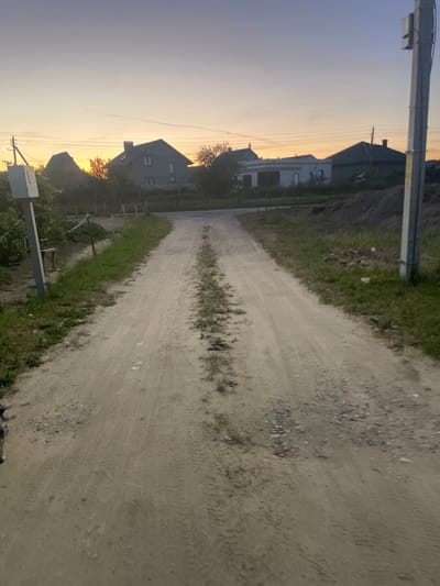 The image depicts a dirt road in a rural setting at dusk. The sun is low on the horizon, casting a warm, golden light that illuminates the sky and hints at the approaching sunset. The road is unpaved and shows tire tracks, suggesting it is used by vehicles. On the left side of the road, several houses with dark roofs and light-colored walls are visible, nestled amongst some trees. The houses appear to be modest residential buildings. The surrounding vegetation is predominantly green grass and some scattered weeds. On the right side of the road, the terrain appears to be slightly elevated and covered with dirt and stones. A utility pole with a box attached to it stands in the foreground on the left side of the image. The overall atmosphere is peaceful and serene, characteristic of a rural landscape during the evening. There is no visible text in the image.