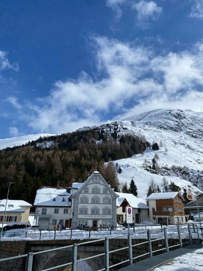 This image depicts a winter scene in Hospental, Switzerland, featuring buildings at the base of a snow-covered mountain under a partly cloudy blue sky.
In the foreground, a metal bridge railing, partially obscured by snow, extends horizontally across the frame. Below the railing, a stone archway of a bridge is visible. The ground surrounding the bridge and buildings is covered in snow.
The central subject is a multi-story building identified as "GASTHAUS ST. GOTTHARD" by a sign on its facade. This building has a light-colored exterior with intricate window frames and a distinctive stepped gable roof, parts of which are covered in snow. To its left, a smaller, white building with a brown roof is visible, also snow-covered. To the right of the Gasthaus, another building with a brown facade and a lower-level garage or entrance is present.
Several indistinct figures are visible on the snow-covered area in front of the Gasthaus, some near tables, suggesting outdoor activity. One individual appears to be holding skis. A blue and white regulatory sign is positioned near the Gasthaus entrance. A dark-colored sedan is parked near the brown-façaded building on the right.
In the midground, behind the buildings, a dense forest of coniferous trees, largely brown due to the season, covers the lower slopes of a mountain. The background is dominated by a large, snow-covered mountain, showing patches of rock and several cleared areas or avalanche barriers on its slopes. The sky above is clear blue with scattered white, wispy clouds. The scene is illuminated by daylight, indicating a sunny winter day.