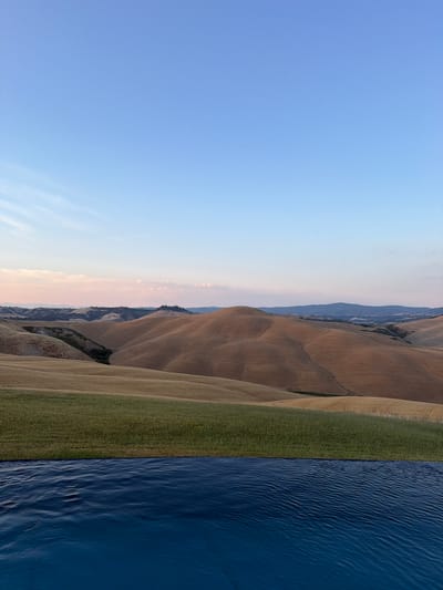The image depicts a serene landscape in Asciano, Italy, with rolling hills and a clear blue sky. In the foreground, the edge of an infinity pool is visible, its deep blue water reflecting the sky and creating a sense of depth. Beyond the pool, a strip of vibrant green grass separates it from the undulating hills, which are covered in dry, golden vegetation, suggesting a late summer or early autumn harvest. The hills are sculpted by the gentle curves of the land, creating a picturesque panorama. In the distance, the landscape continues with more hills, some darker and possibly forested, and a faint haze suggests a sprawling town or village nestled within the terrain. The sky is a gradient of soft blue, with a few wispy, pale pink clouds near the horizon, indicating either early morning or late evening light. The overall atmosphere is tranquil and idyllic.