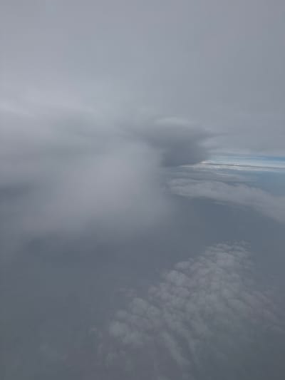 The image depicts a view from above the clouds, likely from an airplane, with the City of Edinburgh, United Kingdom, visible in the distance. The sky is filled with various cloud formations, ranging from dense, white, puffy clouds to wispy, grey layers. The sun is not directly visible, but its light illuminates the scene, suggesting it is daytime, possibly morning or afternoon.
In the mid-ground, a distinct horizon line separates the clouds from the land below. The City of Edinburgh appears as a blurred, distant landscape, hinting at urban structures and geographical features. The clouds themselves are the primary subjects, with different textures and densities creating a dynamic visual. Some areas are brightly lit, indicating thinner cloud cover or direct sunlight, while others are darker and more opaque.
The perspective is from a high altitude, looking downwards and slightly forwards, giving a sense of flight. The clouds are so thick in the foreground that they obscure much of the view, creating a soft, atmospheric effect. The overall impression is one of serene vastness, characteristic of aerial views above the weather. No people or specific objects are discernible in the image, and there is no visible text. The weather appears to be partly cloudy, with the possibility of a bright sky beyond the visible cloud layers. The location context suggests a scene over or near Edinburgh.