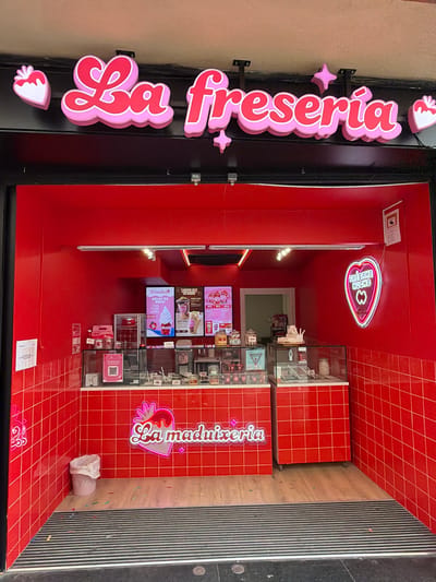 The image shows the storefront of "La freseria", a dessert shop in Granollers, Spain. The shop's exterior is primarily red, with a large pink and red sign on the left side. Inside, the shop has red walls with a grid pattern. Display cases and menu boards are visible, featuring a variety of desserts. A neon sign with a heart and the text "Fresame Mucho" hangs from the ceiling. Another sign, "La maduixeria," is on the right side. There is no person visible in the image.