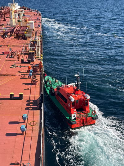 This is an aerial, full shot of a red and green pilot boat approaching the side of a large, red-decked cargo ship. The pilot boat is actively moving, creating a white wake in the dark blue ocean. Several individuals in orange jumpsuits are visible on the deck of the cargo ship, some appearing to be directing the pilot boat. Two individuals are visible on the pilot boat, one standing on the deck and another in the enclosed cabin. The cargo ship's deck is littered with various industrial equipment, including pipes, railings, and bollards. The ocean appears calm with moderate chop. The location is at sea, off the coast of an unspecified location, as no city or landmark is identifiable in the image.