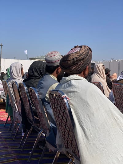 This image depicts an outdoor gathering in Mandisar, Afghanistan. A line of individuals, primarily male, are seated in chrome-framed chairs with patterned maroon and black fabric backs, arranged on a striped mat of blue, purple, red, and yellow. They are viewed from behind, facing an unseen focal point. The closest individual wears a dark brown turban with a central circular emblem and a white garment draped over his back. To his left, another man wears a grey embroidered skullcap with red accents and a light blue vest. Further left, individuals are visible wearing a black patterned head covering and a light-colored head covering, respectively. In the background, a white wall topped with razor wire extends horizontally. Behind this wall, a light-colored building is partially visible, and a white flag, possibly bearing a dark emblem, flies from a pole against a clear blue sky. The scene is under bright daylight.
