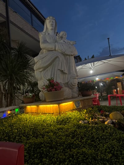 The image captures a twilight scene in Ardea, Italy, focusing on a white marble statue of the Virgin Mary holding the infant Jesus. The statue stands on a raised platform illuminated by warm yellow lights. Surrounding the statue is lush green foliage. To the left, a building with a brick facade and large windows is visible, along with a palm-like plant. To the right, a white umbrella covers an outdoor seating area with red chairs and tables, suggesting a restaurant or cafe. The sky is a deep blue, indicating the transition from day to night. There are no discernible people actively engaged in any specific action within the frame of the main subject.