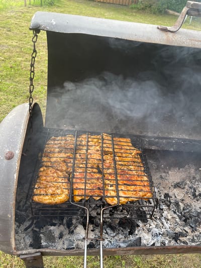 In Kalnciems, Latvia, a barbecue grill is open, with chicken pieces cooking on a wire rack over hot coals. Smoke rises from the grill, and the surrounding area is covered in grass. The grill is made of metal and appears to be old, with some rust and wear. The chicken is golden brown and appears to be seasoned with herbs. The scene is casual and suggests an outdoor meal.