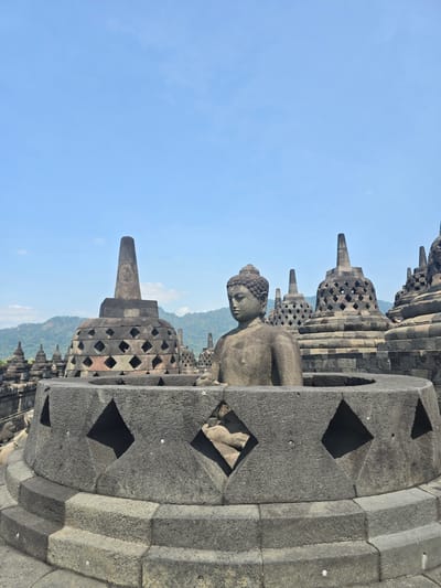 The image depicts a stone statue of a Buddha positioned behind a lattice structure, set against a backdrop of stupas under a blue sky. The scene is part of Borobudur Temple, located in Desa Borobudur, Indonesia. The statue is weathered and seated, and the lattice structure surrounding it has diamond-shaped openings. The stupas in the background are similar in style to the lattice structure, and the landscape includes a glimpse of mountains or hills in the distance.