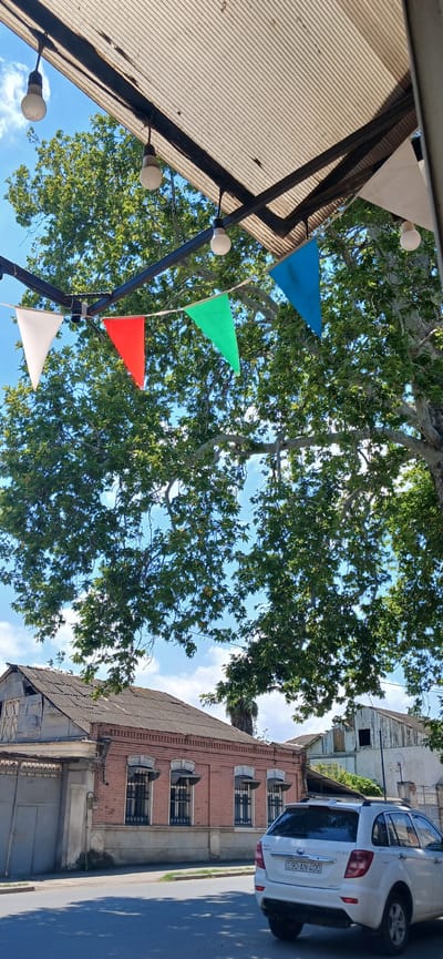 In Ganja City, Azerbaijan, an outdoor scene unfolds beneath a corrugated metal awning, strung with colorful triangular flags and bare light bulbs. These flags, in white, red, green, and blue, hang suspended against a backdrop of a clear blue sky dotted with white clouds, and the lush green foliage of a large tree. Below the awning and tree, an old, weathered brick building with a corrugated roof is visible, featuring barred windows. To its right, a white car, a hatchback with license plate 90-AN-400, is parked on an asphalt road, casting a shadow on the ground. The overall impression is one of a casual, perhaps festive, outdoor setting in a historic urban environment.