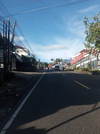 The image displays a paved asphalt road in Khouathineung, Laos, under a clear blue sky with minimal white clouds. The perspective is a low-angle view looking down the road, which features a solid white line on its left edge and a dashed yellow line further right, marking lanes. Long, dark shadows extend across the foreground roadway from objects positioned to the left.
On the left side of the road, a series of utility poles are visible, heavily laden with a dense network of black electrical and communication cables that extend across the upper portion of the frame. Behind these poles, various buildings, including a multi-story white structure, line the street. On the right side, a decorative stone wall with patterned white and dark sections, adorned with ornate pillars, runs parallel to the road. Beyond this wall, buildings include one with a prominent red-orange facade. Further down the road on the right, a large blue sign with white lettering clearly displays the words "BIRD HOTEL".
Traffic is present on the road in the mid-distance: a dark-colored SUV is visible in the right lane, and at least two motorcycles are also present, one positioned ahead of the SUV and another partially obscured on the far right. Several indistinct human figures are discernible on the left side of the road in the far distance, near the buildings, indicating pedestrian presence. The overall scene depicts a daytime urban street environment.