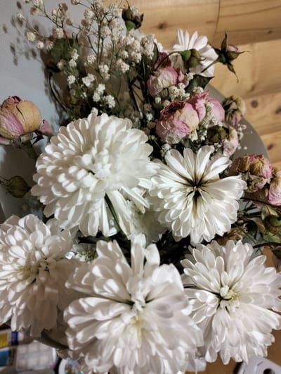 The image shows a close-up view of a bouquet of flowers, predominantly white chrysanthemums, along with dried pink roses and sprigs of baby's breath. The setting appears to be indoors, with a light blue wall visible in the upper left corner and a wooden panel in the background. The specific location in Krykhivtsi, Ukraine cannot be determined from this image.