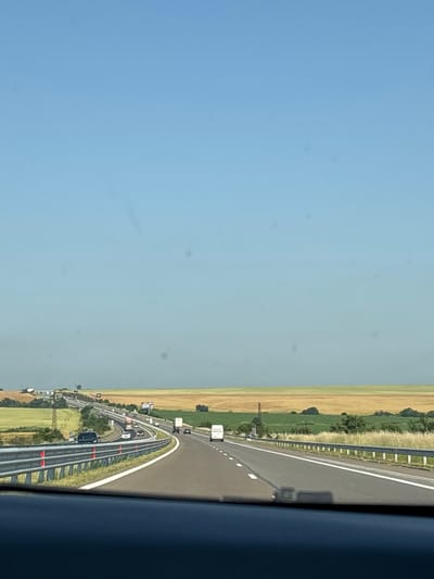 The image displays a multi-lane highway under a clear blue sky, observed from the interior of a vehicle, indicated by a dark dashboard occupying the bottom portion of the frame. Circular dust spots are visible across the sky area.

The highway, appearing to be an expressway or motorway, curves gently to the right in the foreground and features multiple lanes with distinct white markings. Metal guardrails, equipped with red reflectors, line the immediate edges of the road. Moderate vehicle traffic is present: a dark-colored SUV occupies an inner lane in the foreground, while on a higher, parallel section of the highway further to the left, multiple additional vehicles, including a red-cab truck and a white box truck, are visible. A white courier van displaying "ЕКОНТ" (EKONT) branding on its rear is positioned in the right-most lane in the mid-ground.

The surrounding landscape consists of extensive agricultural fields. Fields to the left of the highway are predominantly green, while those to the right exhibit a golden-yellow coloration. Sparse trees and bushes are scattered across the terrain. A metal lattice transmission tower is prominent in the green fields on the left side of the highway. Two billboards are also visible adjacent to the elevated road section on the left. The scene is brightly lit by daylight, indicating clear weather conditions in the Hadzhiite area of Bulgaria.