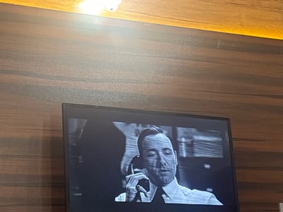 A television screen displays a black and white image of a man in a suit and tie holding a vintage telephone. The television is mounted on a wood-paneled wall. The scene appears to be indoors. The location, Eneka, Nigeria, cannot be determined from the image.